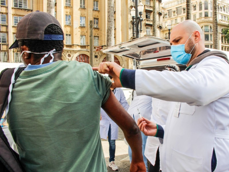 Agente de saúde do Consultório na Rua aplica uma vacina em uma pessoa em situação de rua, que está de costas
